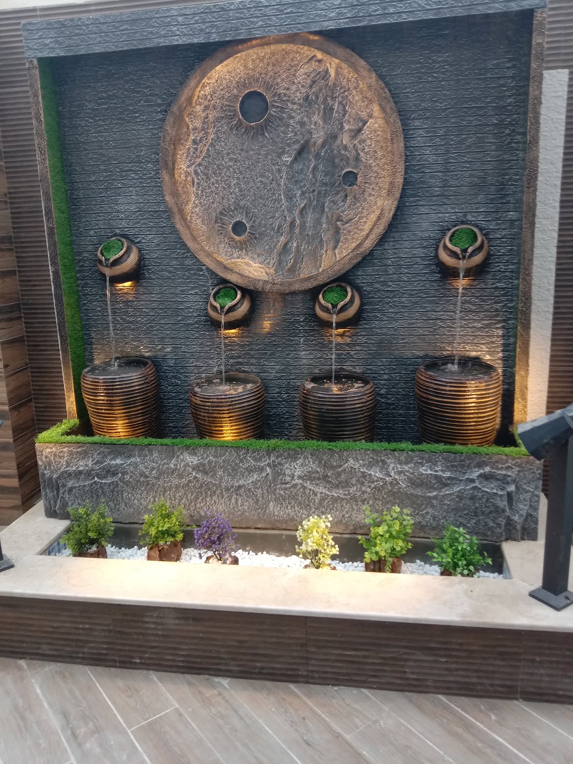 A decorative wall-mounted water feature with a large circular bronze-toned sculpture on a dark textured backdrop, five tiered metal basins with green stone accents, small potted plants along the front ledge, and warm under-lighting.