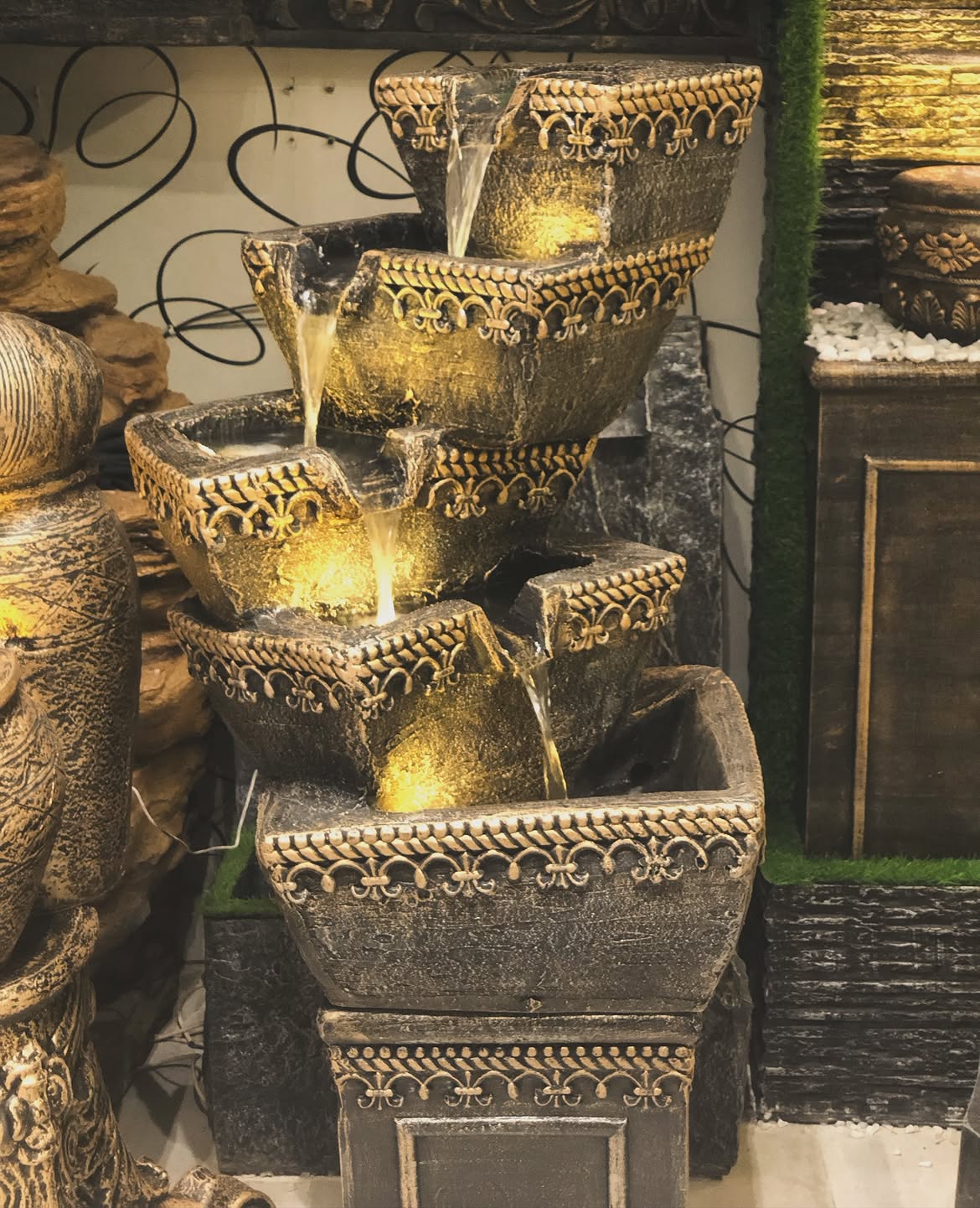 A tiered outdoor water fountain with ornate gold and dark gray stone-like detailing, illuminated by warm yellow underlight, shown in a garden or patio setting.
